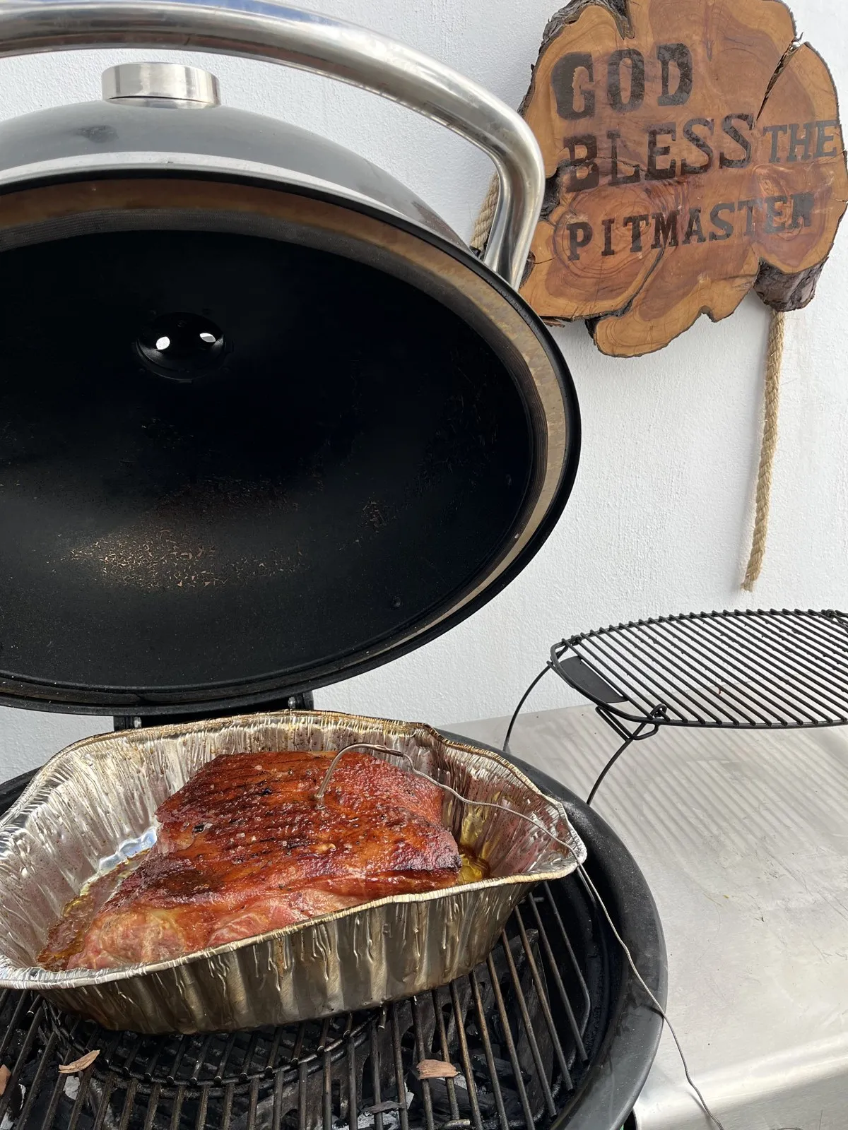 Paleta de cerdo con costra de rub en el Kamado abierto, con el letrero God Bless the Pitmaster atrás